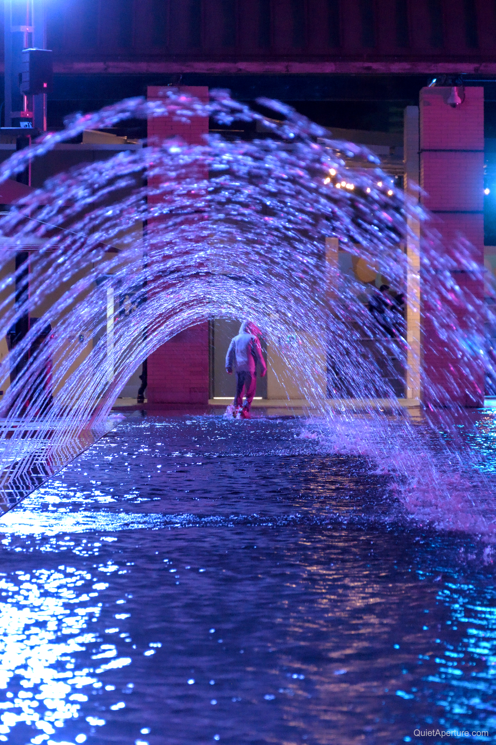 Blue and violet fountain arches framing a person at night, frame 03.